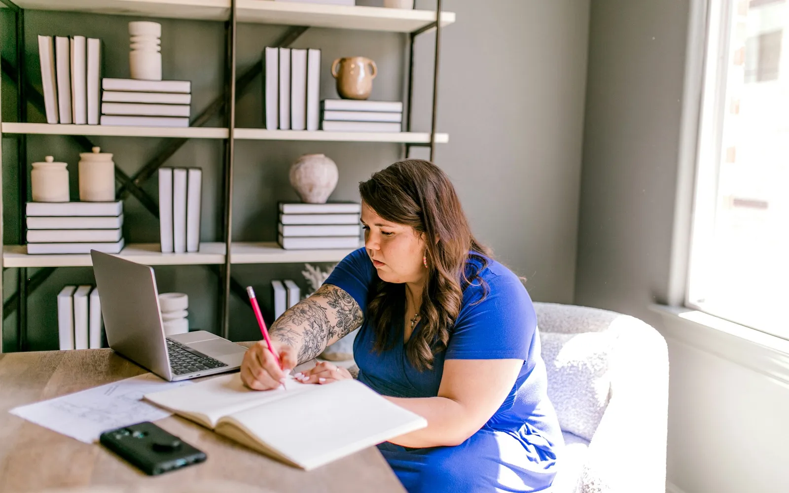 Small business owner working through numbers in a notebook beside a laptop at a desk by the window