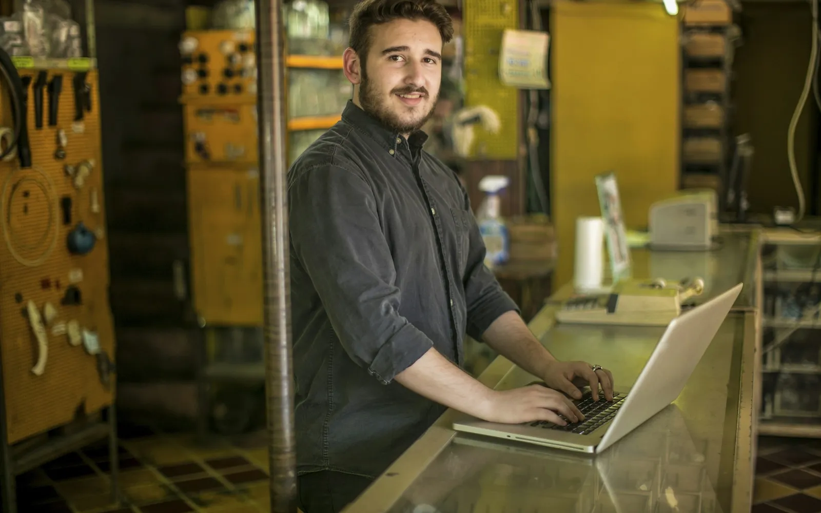 Shop owner standing at the glass counter of a parts store typing on a laptop