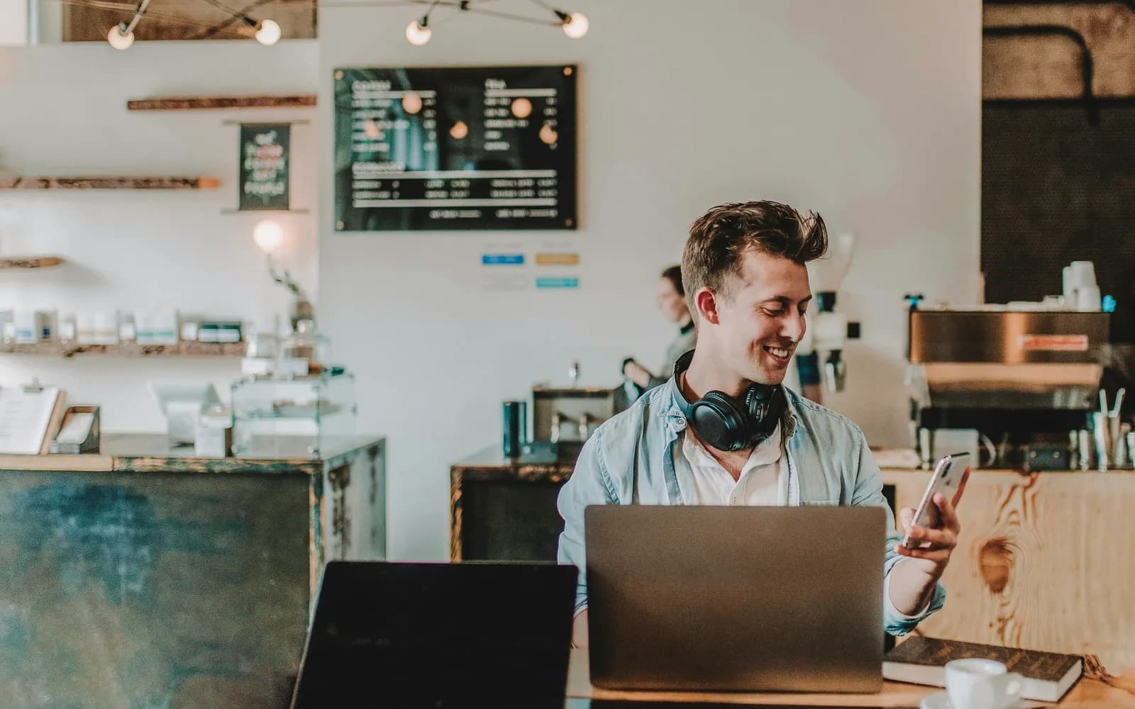 Cafe owner smiling at a phone with a laptop open at a table near the counter