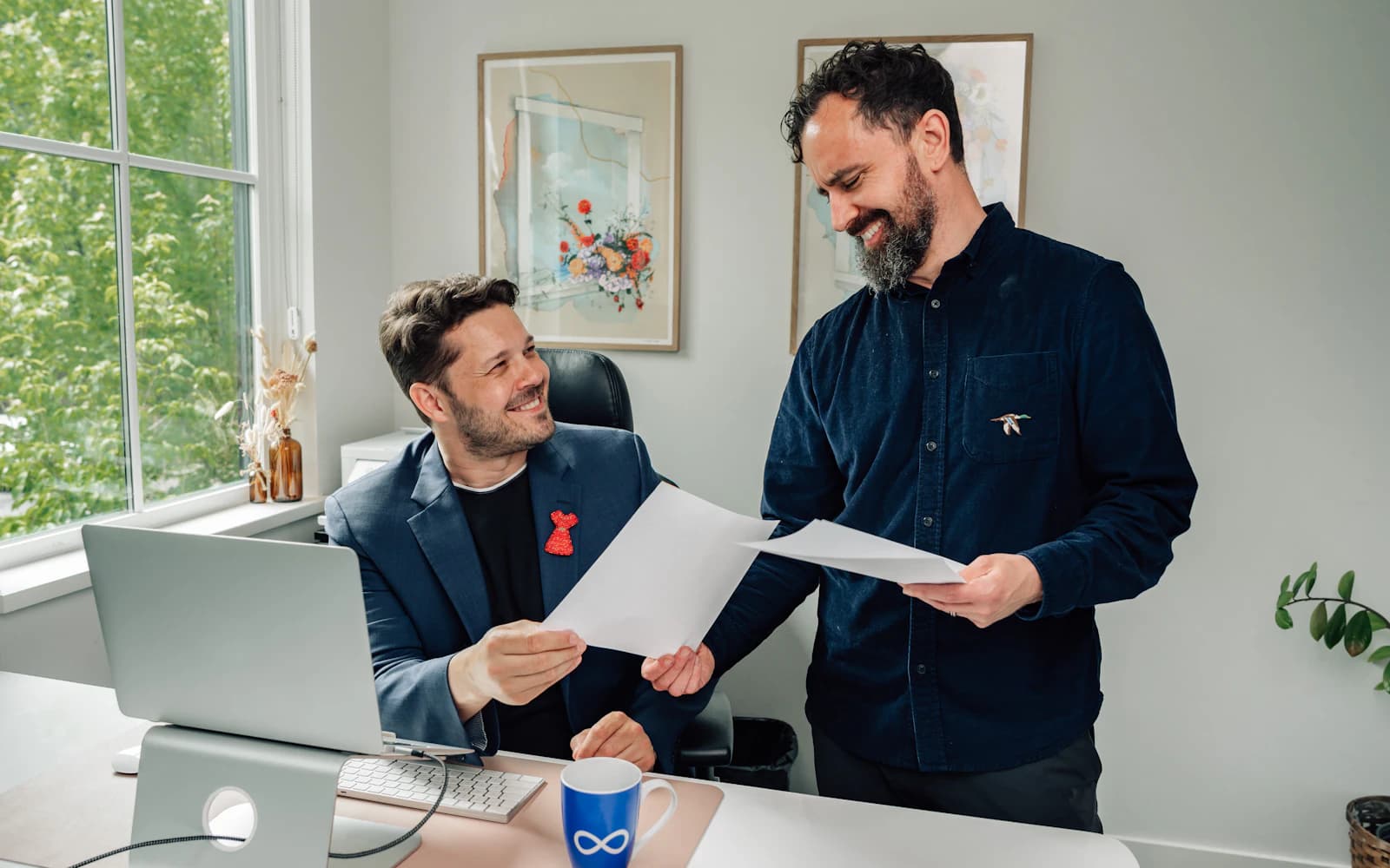 Two business owners comparing printed documents at a desk beside a laptop