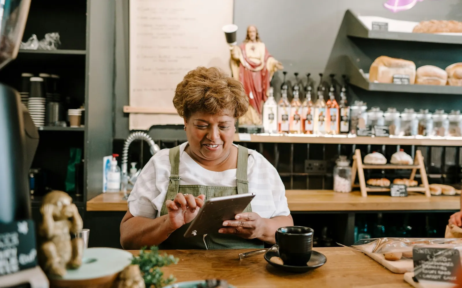 Cafe owner in an apron checking sales on a tablet at the counter beside a cup of coffee
