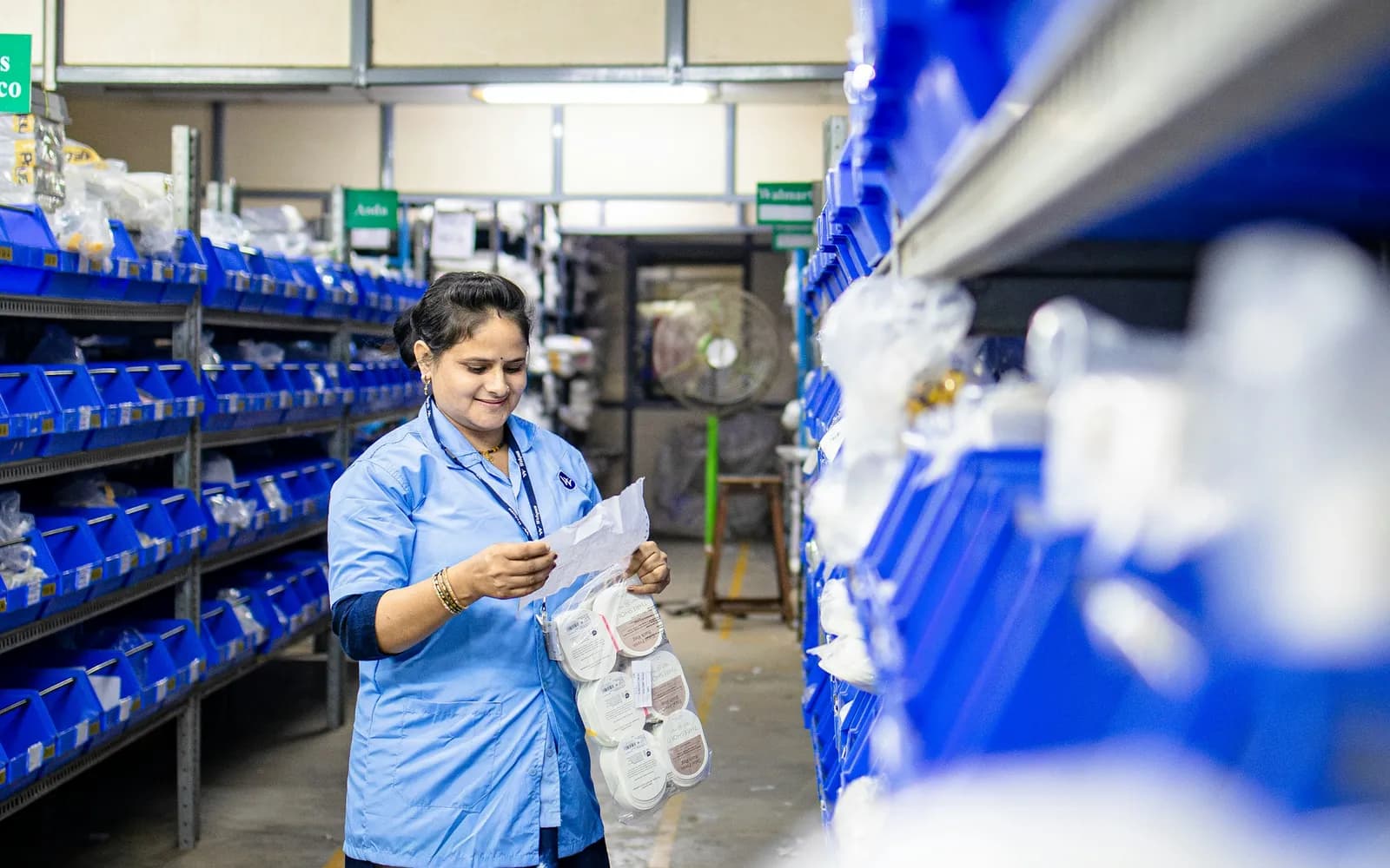 Warehouse owner checking stock against a printed list between shelves of blue bins