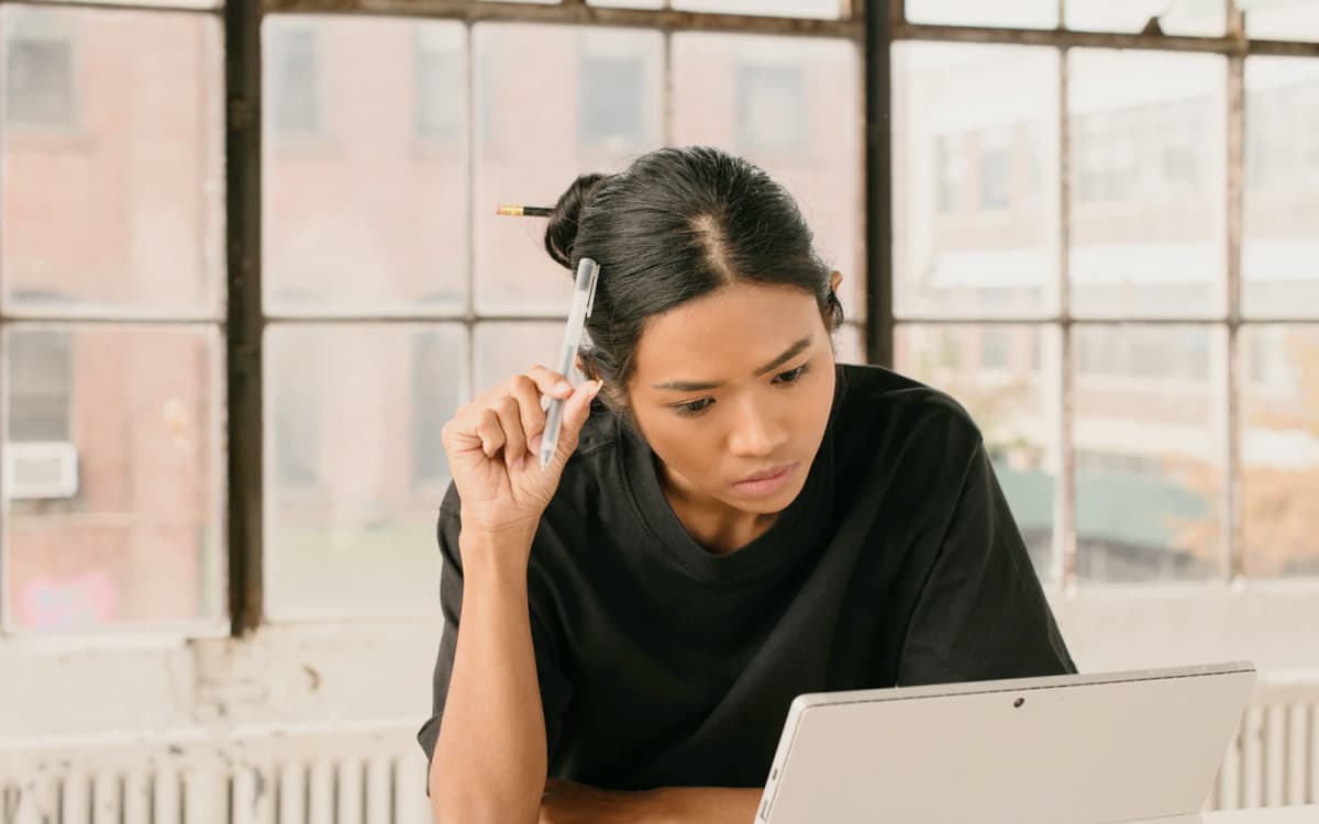 Business owner with a pen in hand reading her laptop screen at a table by the window