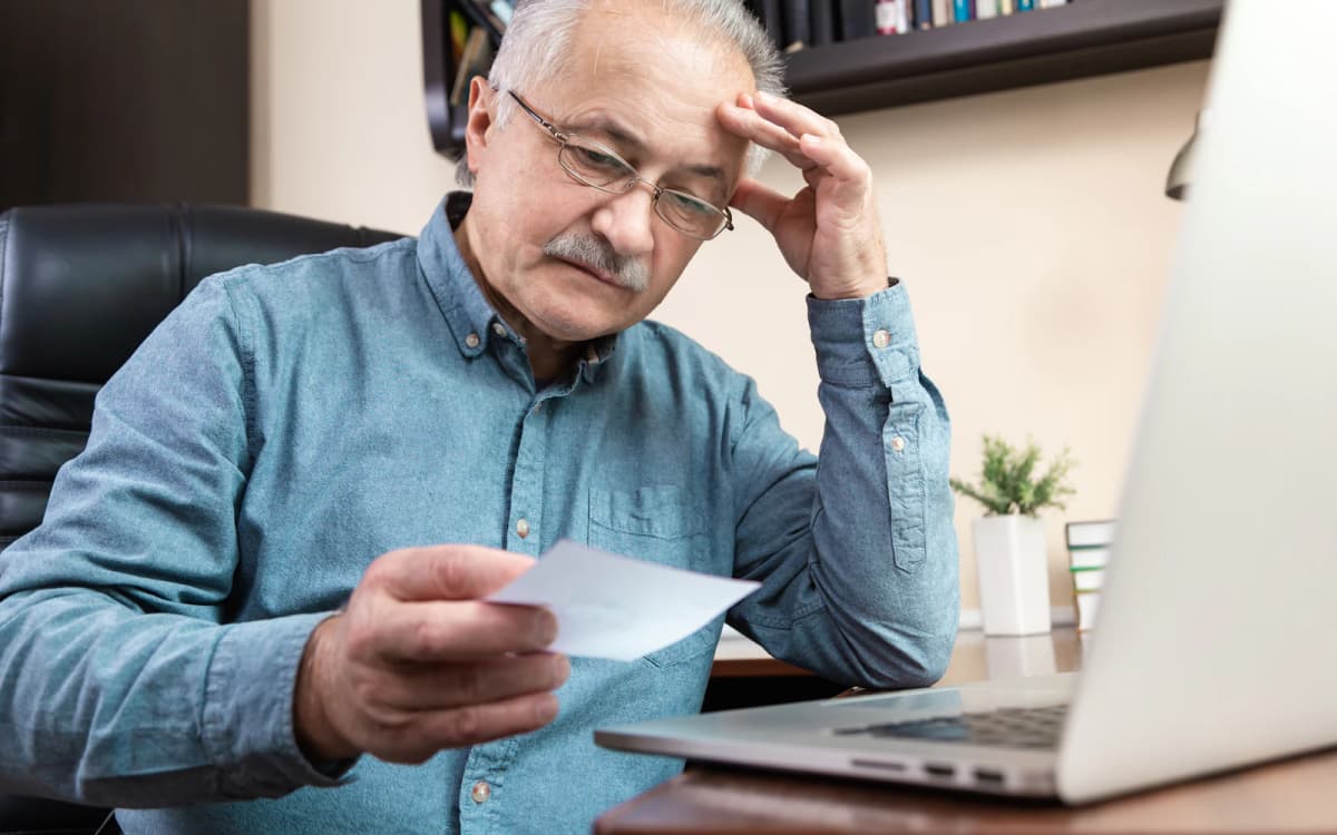 Older business owner looking over a paper slip beside his laptop with a worried expression