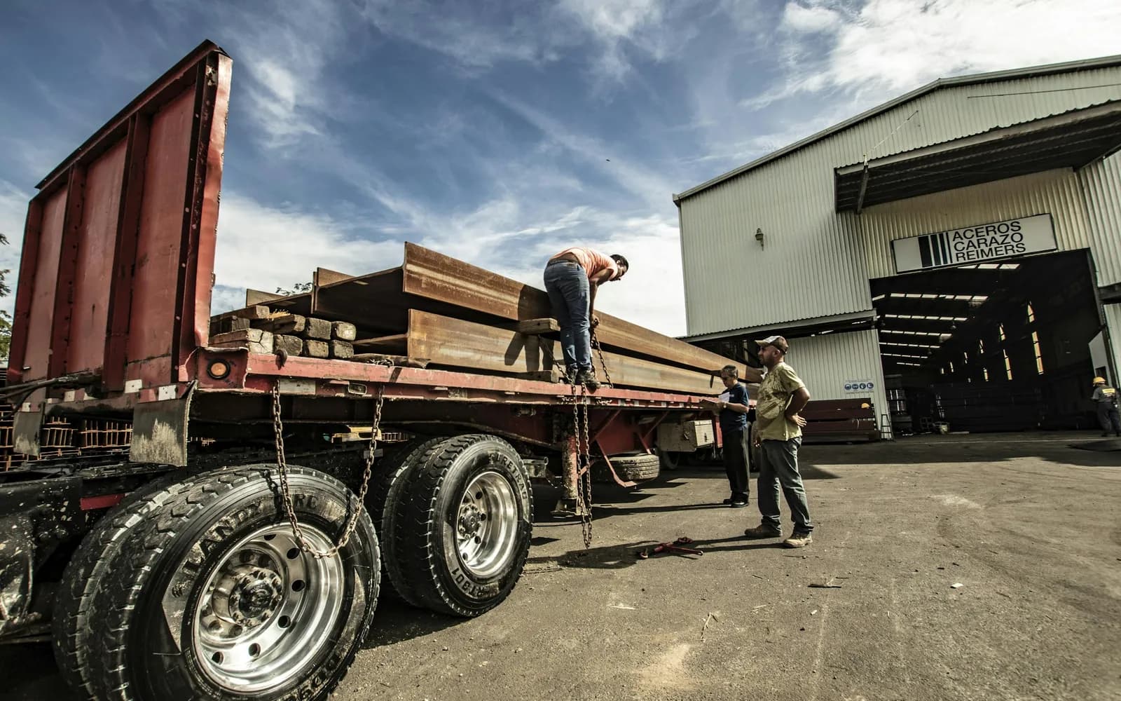 Crew unloading steel beams from a flatbed truck outside a supply warehouse