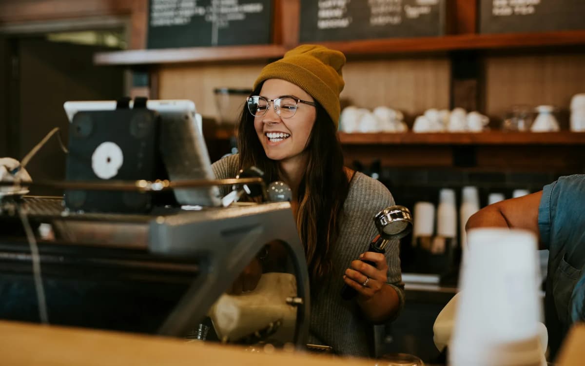 Smiling coffee shop owner in glasses and a beanie behind the counter next to a tablet register
