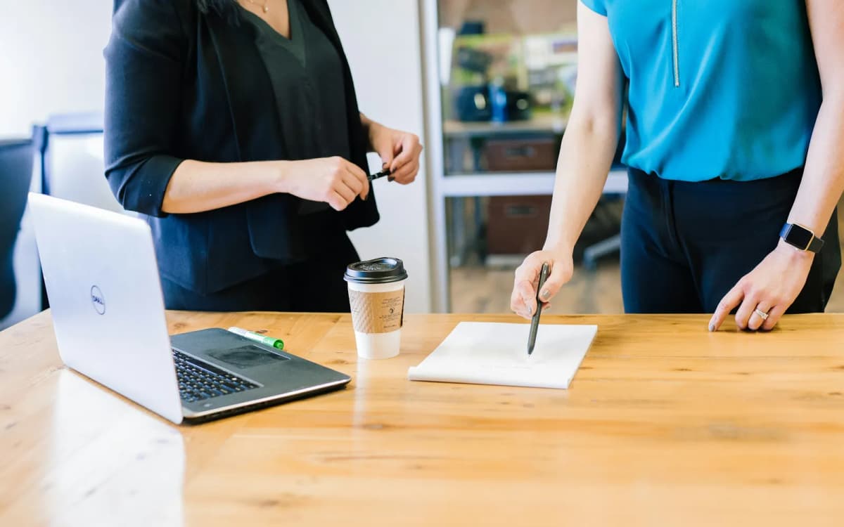 Two colleagues at a wooden table checking a printed page against figures on a laptop