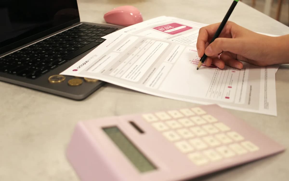 Hand marking figures on a printed statement beside a laptop and a pink calculator