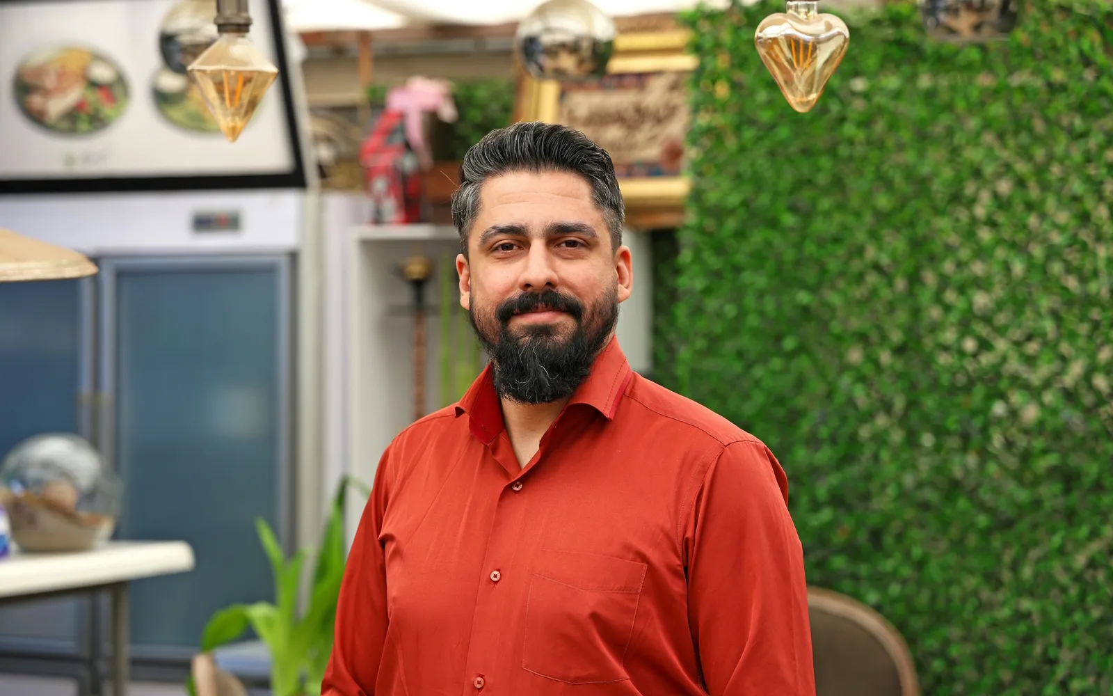 Smiling restaurant owner standing inside a small dining room beside a green plant wall