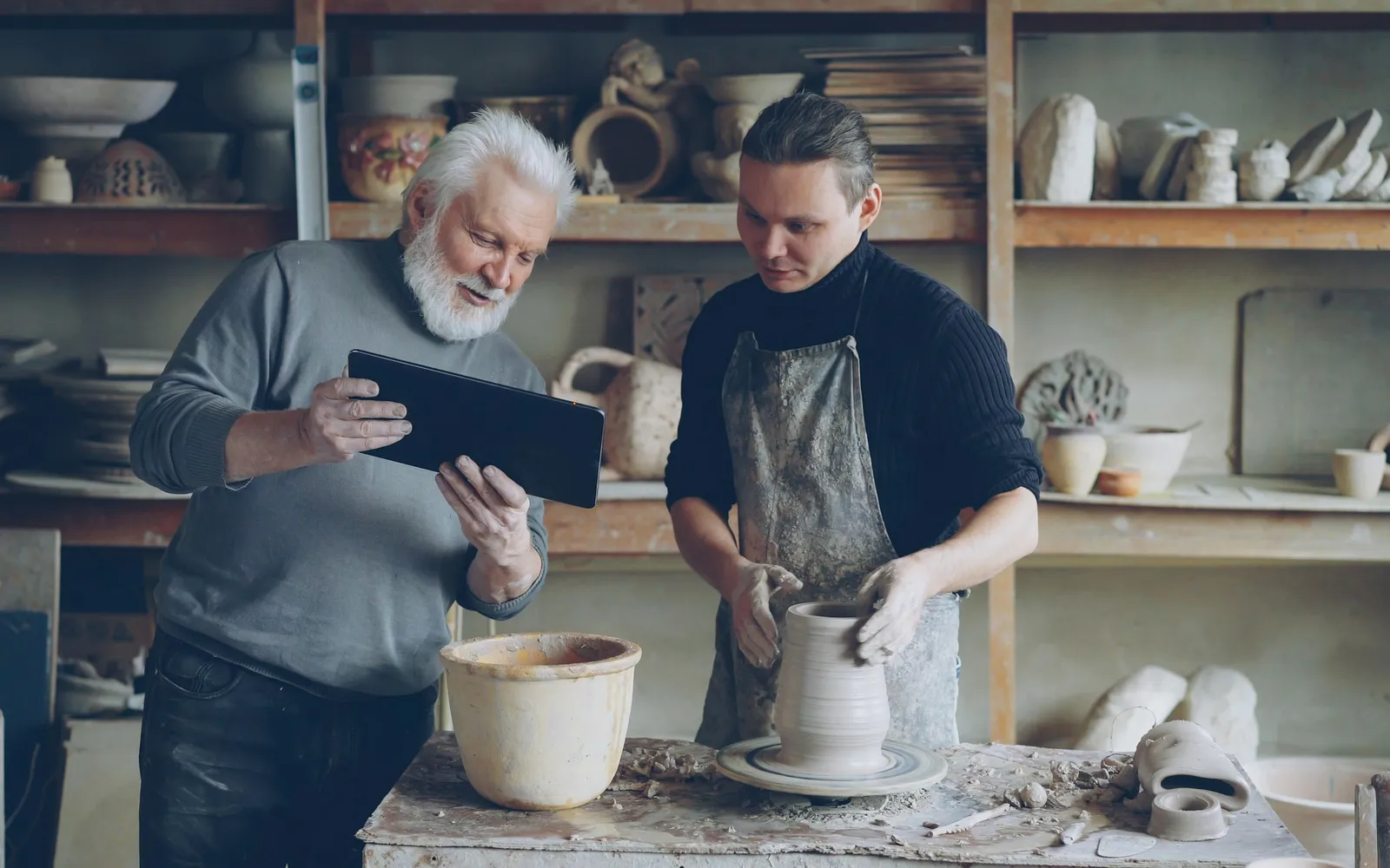 Older pottery studio owner showing a tablet to a younger potter working at the wheel