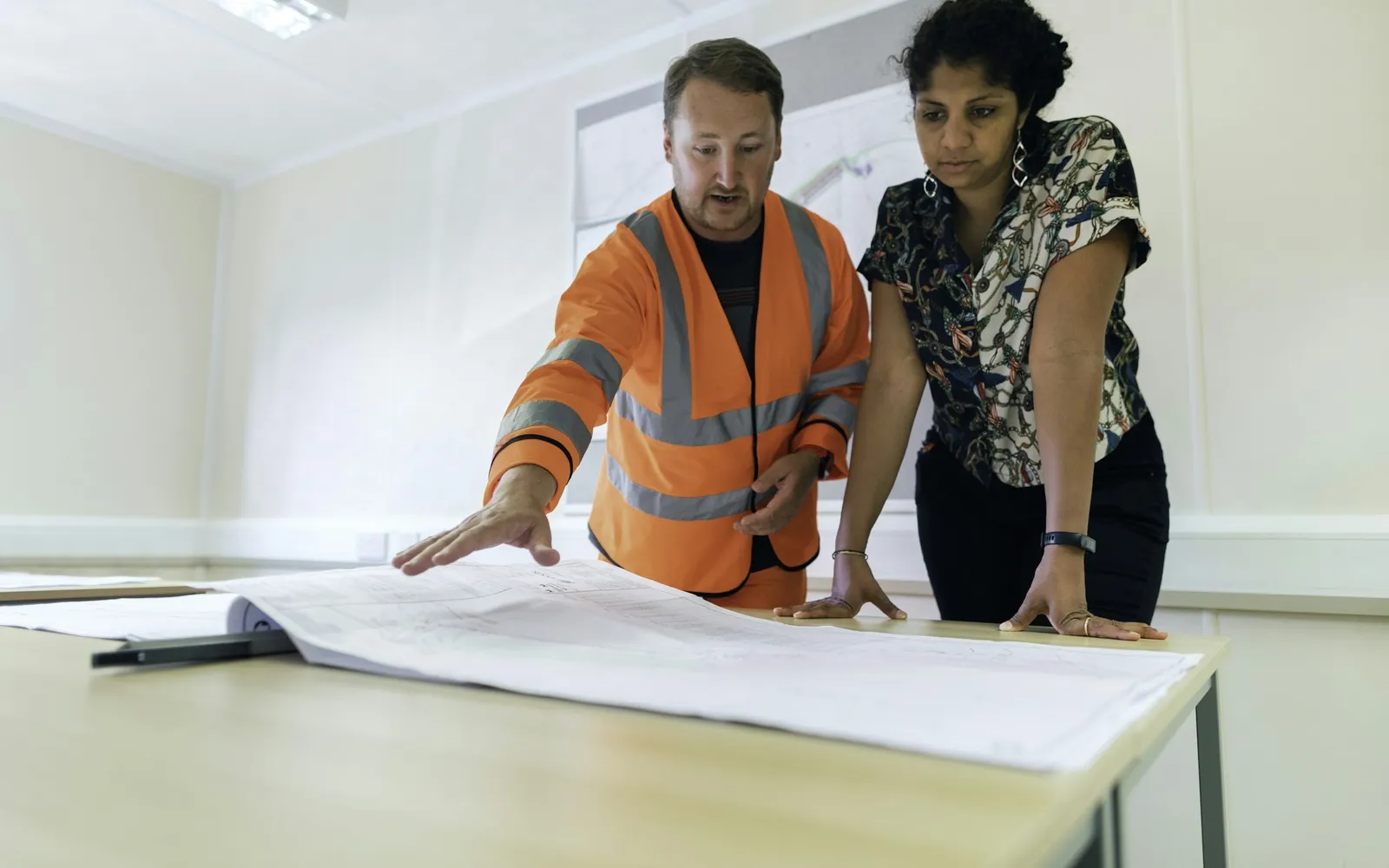 Contractor in a high-visibility vest reviewing plans with a colleague at a site office table