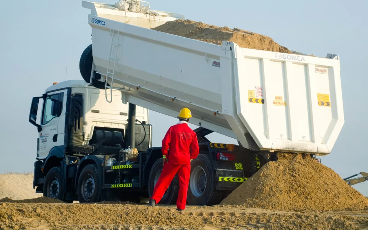 Site worker in red coveralls watching a dump truck unload sand at a work yard